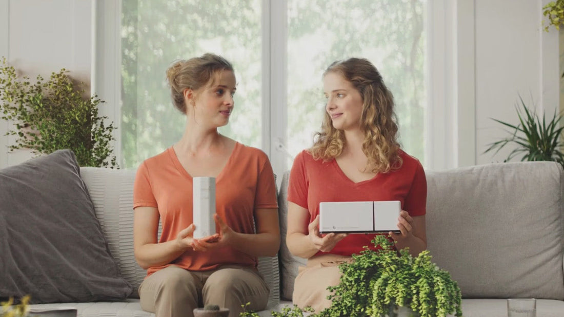 Two women sitting on a couch in a bright room with green plants surrounding them, holding probiotic machines