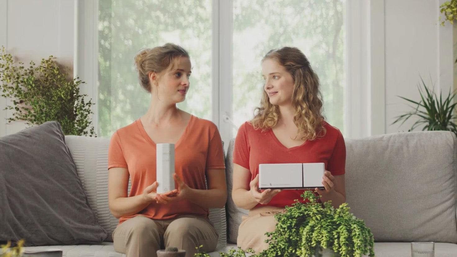 Two women sitting on a couch in a bright room with green plants surrounding them, holding probiotic machines
