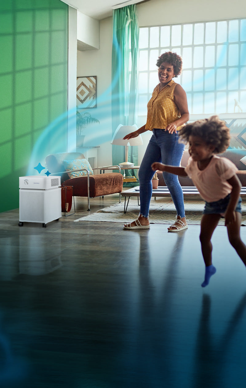 Woman and child playing in a modern living room with green walls and large windows, an air purifier sits in the corner with blue wavy air lines emanating from it