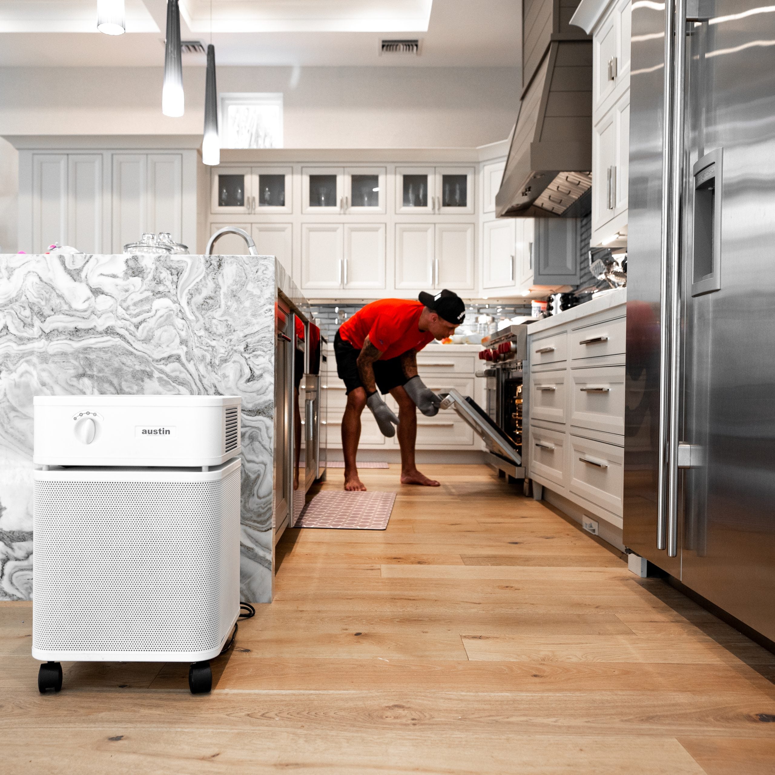 Modern kitchen with a person using an oven, a white Austin air purifier on the floor, and stainless steel appliances.