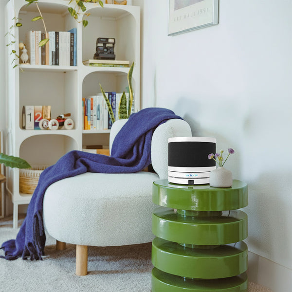 Green side table with a cylindrical Air Purifier next to a white armchair with a blue blanket in a room with a bookshelf