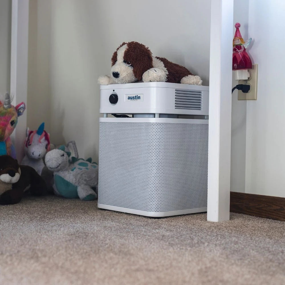 White air purifier with a plush dog toy on top, surrounded by stuffed animals in a room.